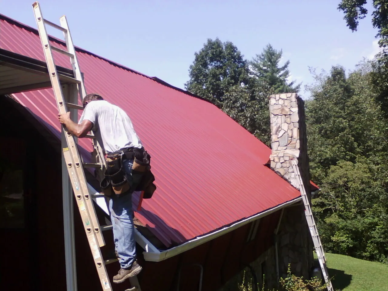 Roofer on a ladder beside a red metal roof and stone chimney — Soffit & Fascia Repair by Roofing Brothers in Souderton